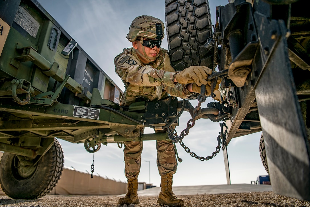 A soldier reaches to connect a trailer to the back of a Humvee using a chain.