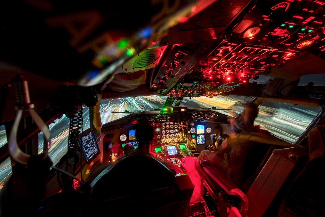 Airmen sit in a cockpit illuminated by red, green and blue lights.