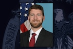 Head and shoulders image of a white man in a black suit jacket, white shirt and red tie posing in front of the American flag.