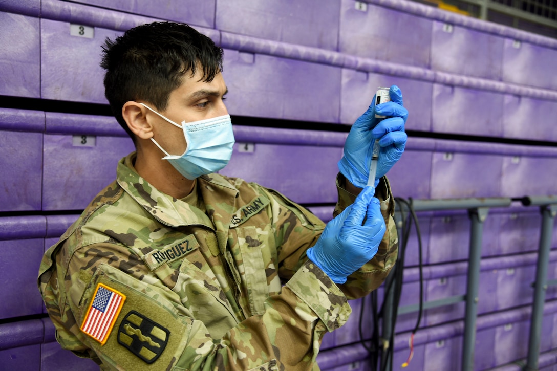 A soldier wearing a mask holds up a COVID-19 vaccine vial and syringe.