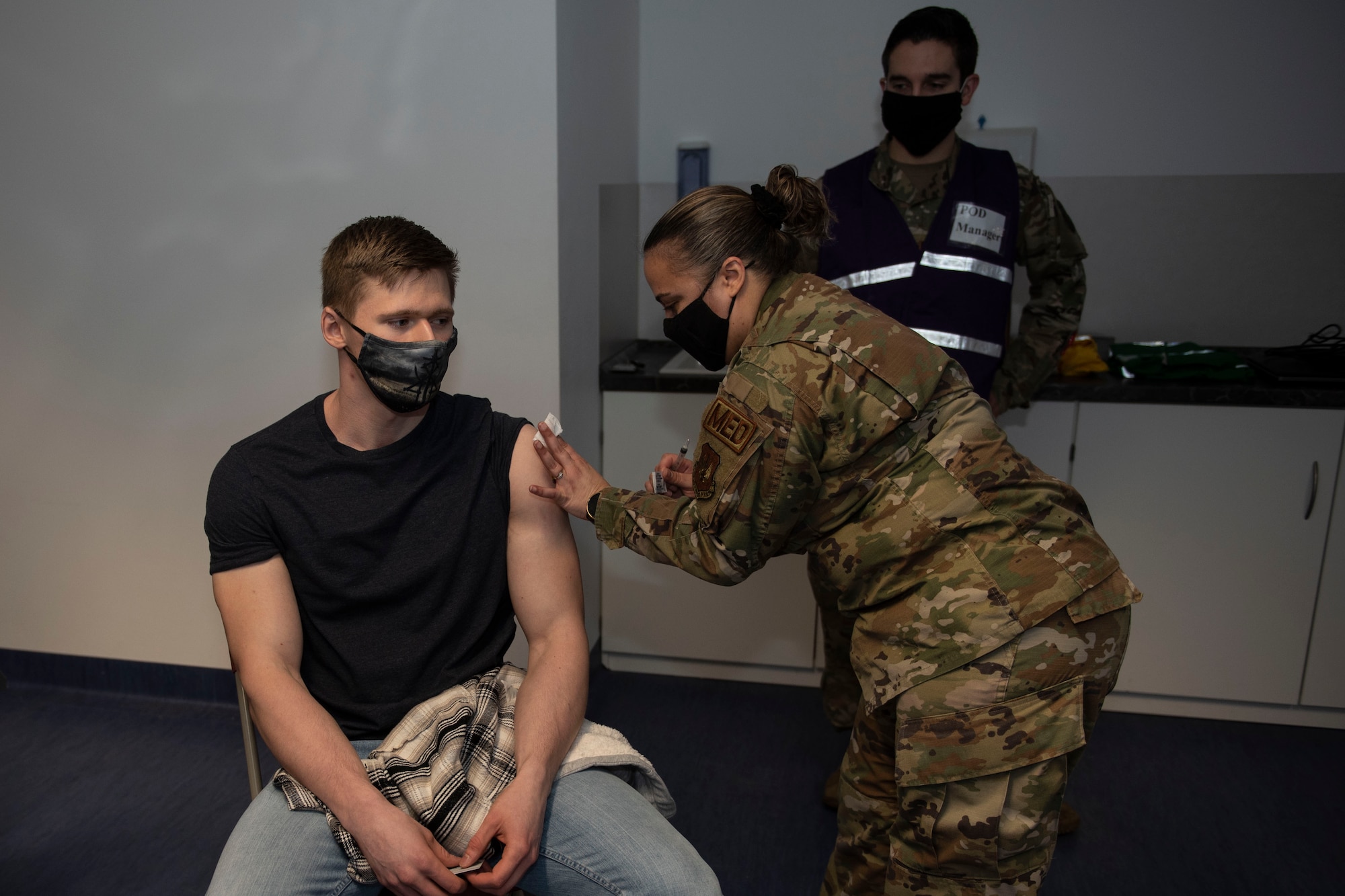 U.S. Air Force Senior Airman Brandon Franklin, 52nd Civil Engineer Squadron Fire and Emergency Services firefighter, receives the COVID-19 vaccine from Tech Sgt. Valeria Feist, 52nd Medical Operations Squadron allergy and immunization technician, Jan. 4, 2021, at Spangdahlem Air Base, Germany. Capt. Matthew Jordan, 52nd Medical Support Squadron chief of pharmacy operations, back, oversaw and hosted the first distribution of COVID-19 vaccinations for 52nd Fighter Wing Airmen, and ensured the process and comfort of service members were a priority. (U.S Air Force photo by Senior Airman Melody W. Howley)