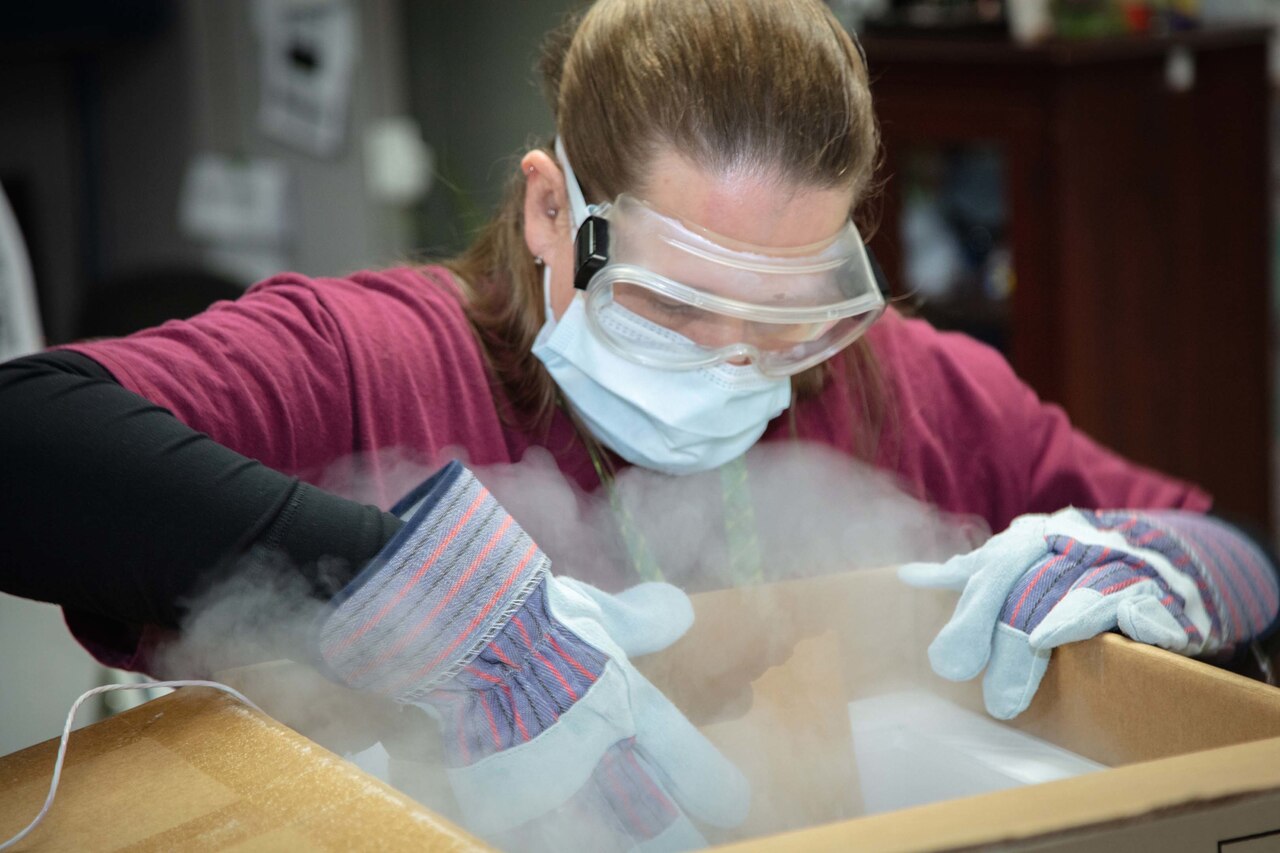 A woman wearing eye goggles, mask and gloves takes vaccine out of a box.