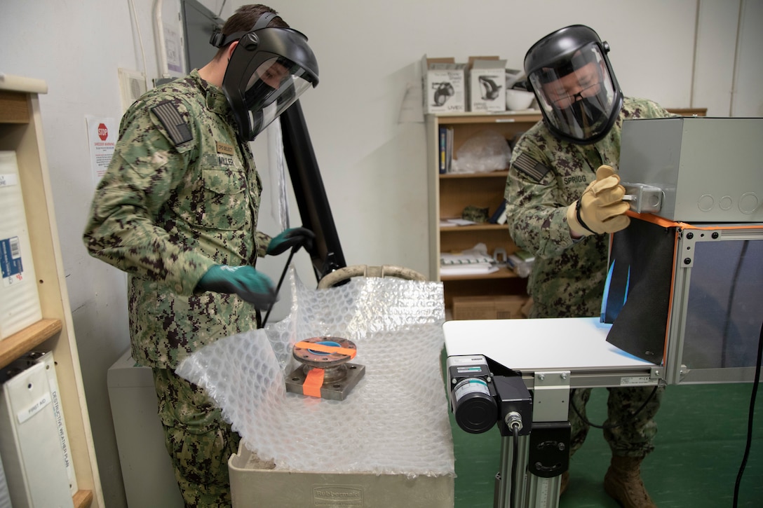 A service member wraps something in bubble wrap while another holds a square metal object.