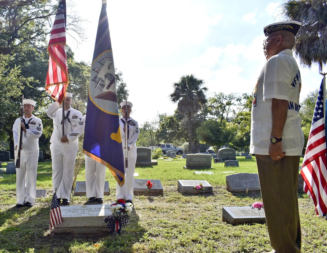 A man salutes a color guard standing in front of a gravesite in a cemetery.