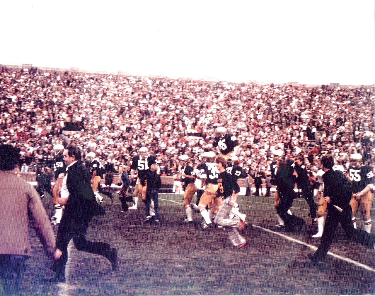 People are running off a football field as a football player is carried by teammates on their shoulders. In the background, the stands are filled with people.