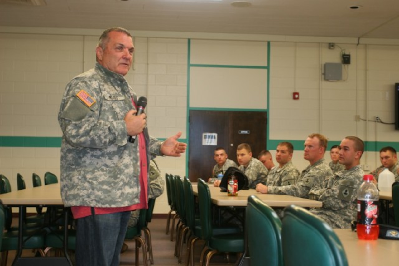 A man speaks into a microphone as soldiers sit at tables to listen.