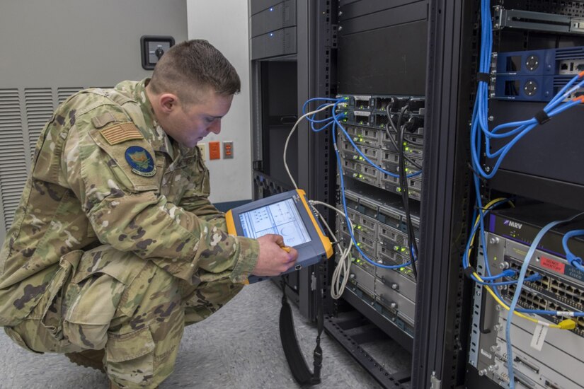A man dressed in a military uniform squats on the floor to look at a device he holds in his hands; electrical equipment is in front of him.