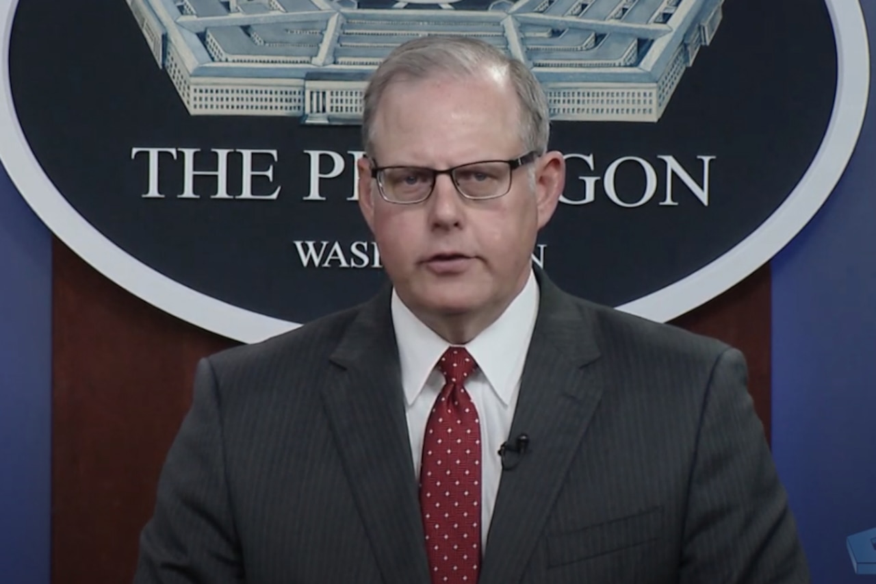 A man dressed in a business suit looks forward into a camera and speaks.