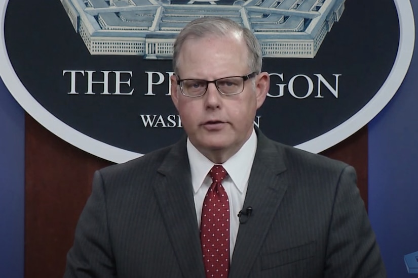 A man dressed in a business suit looks forward into a camera and speaks.