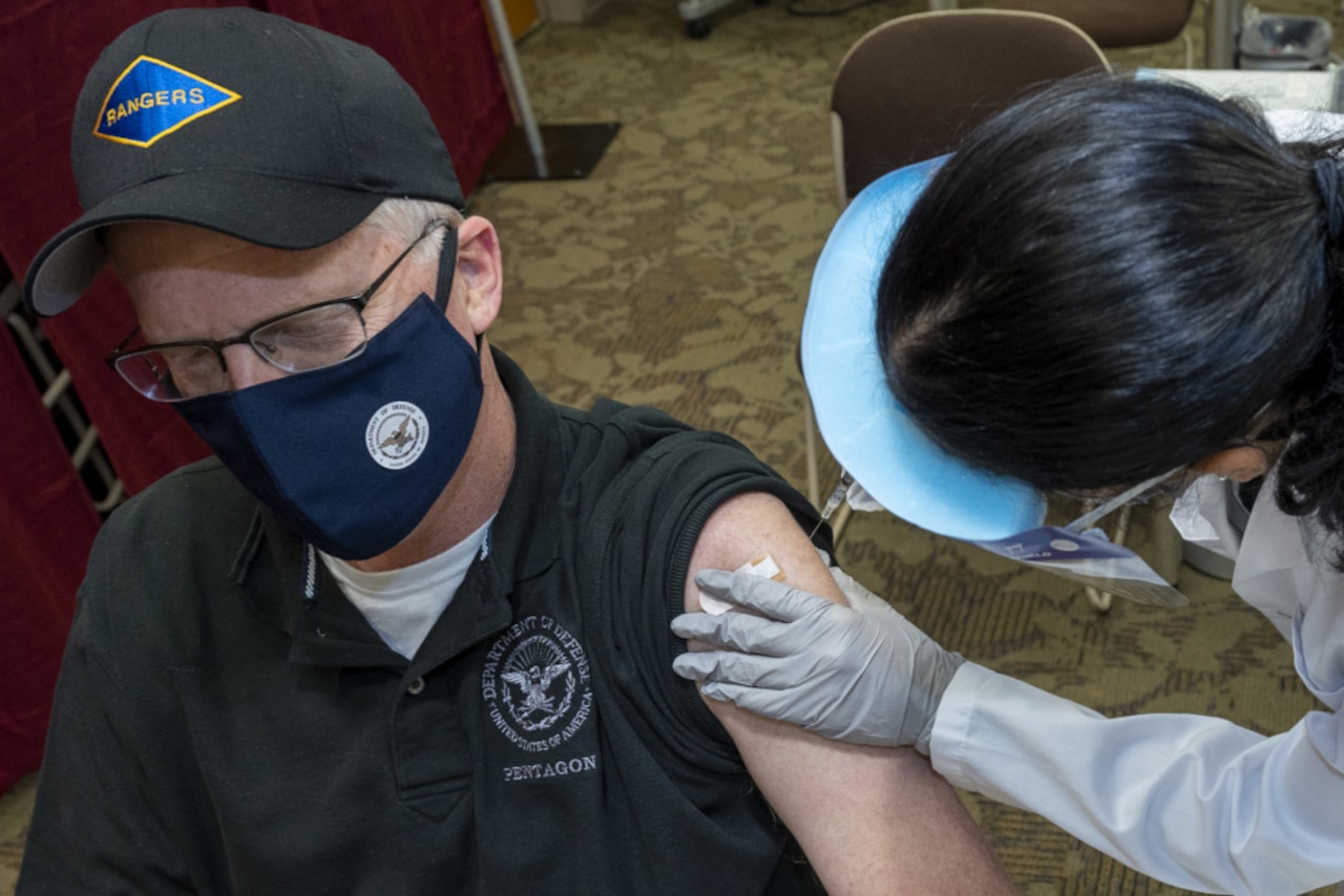A man wearing a mask sits while a medical technician gives him an injection in the arm.