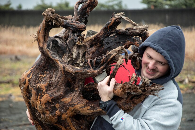A Marine in civilian clothes carries what looks like a large piece of driftwood on a beach.