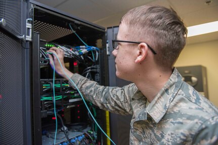 A young man dressed in a military uniform reaches into a cabinet with electrical equipment.