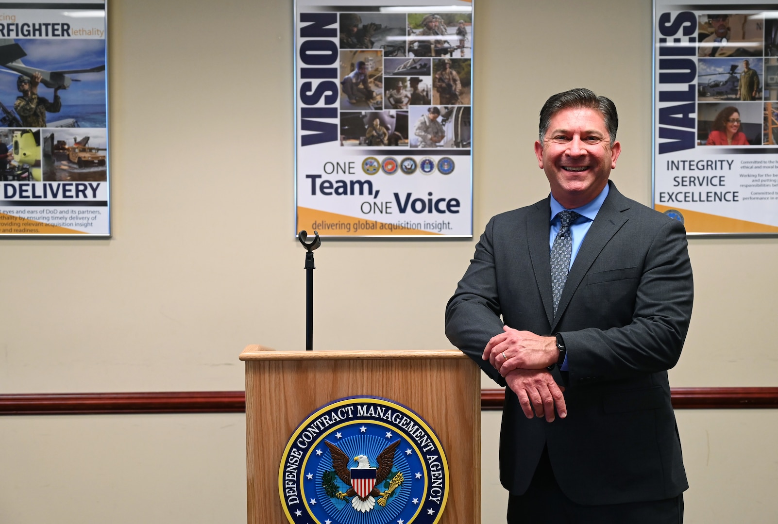 Man wearing a suit and tie stands besides a podium with the DCMA framed posters in the background