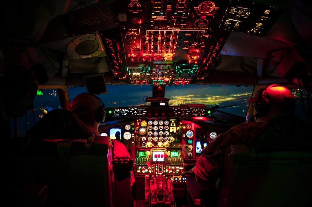 Two pilots sit in a cockpit illuminated by red light.