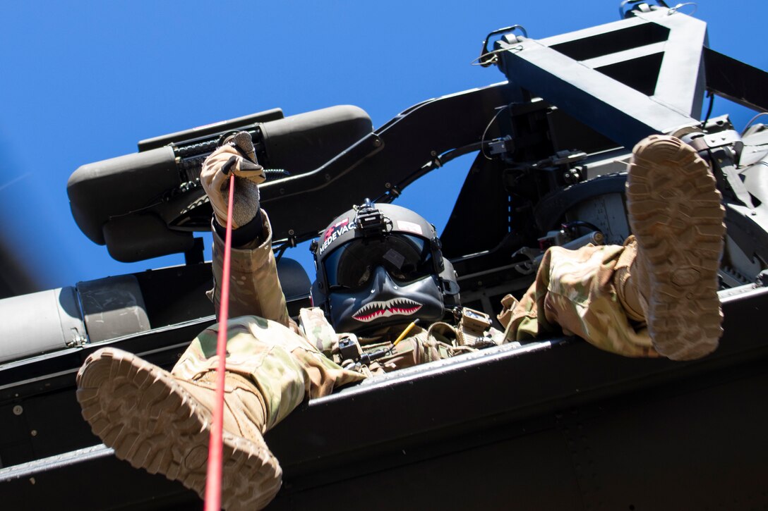 A soldier wearing a mask looks down from a helicopter while holding a rope.