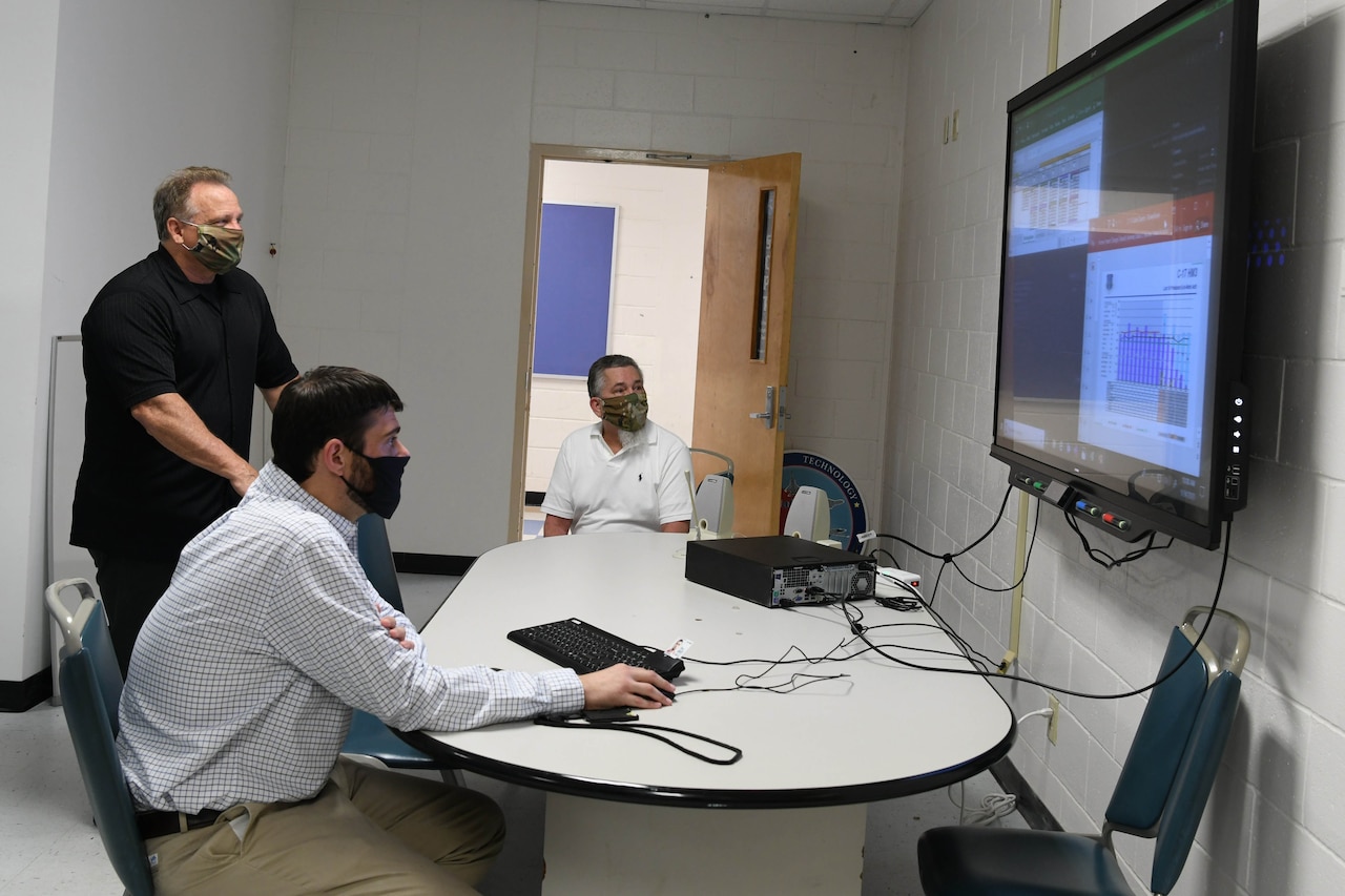 Three men -- two seated and one standing -- gather around a table and look at a giant computer screen hanging on a wall. A computer and keyboard sit on the table.