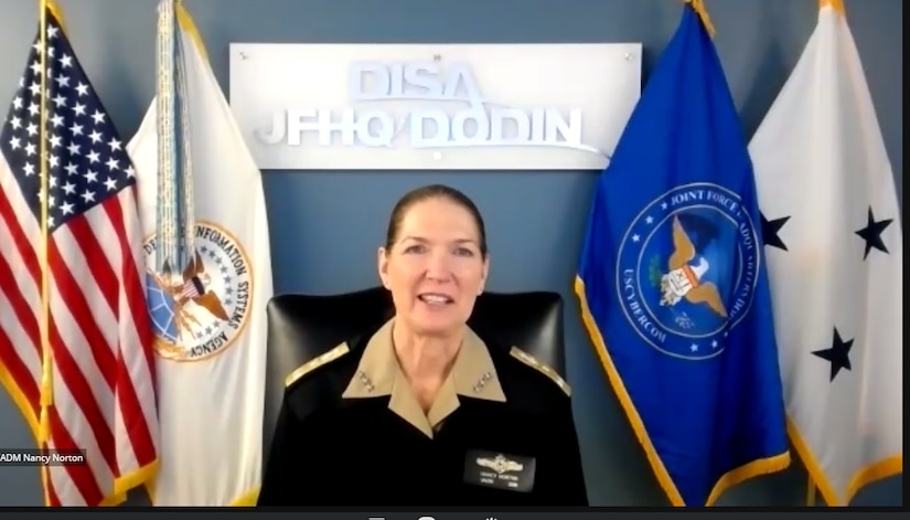 A woman in an officer’s uniform, flanked by flags, sits in a chair and looks forward as she speaks to a camera.