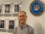 A man stands in an office hallway with a wall of portraits behind him