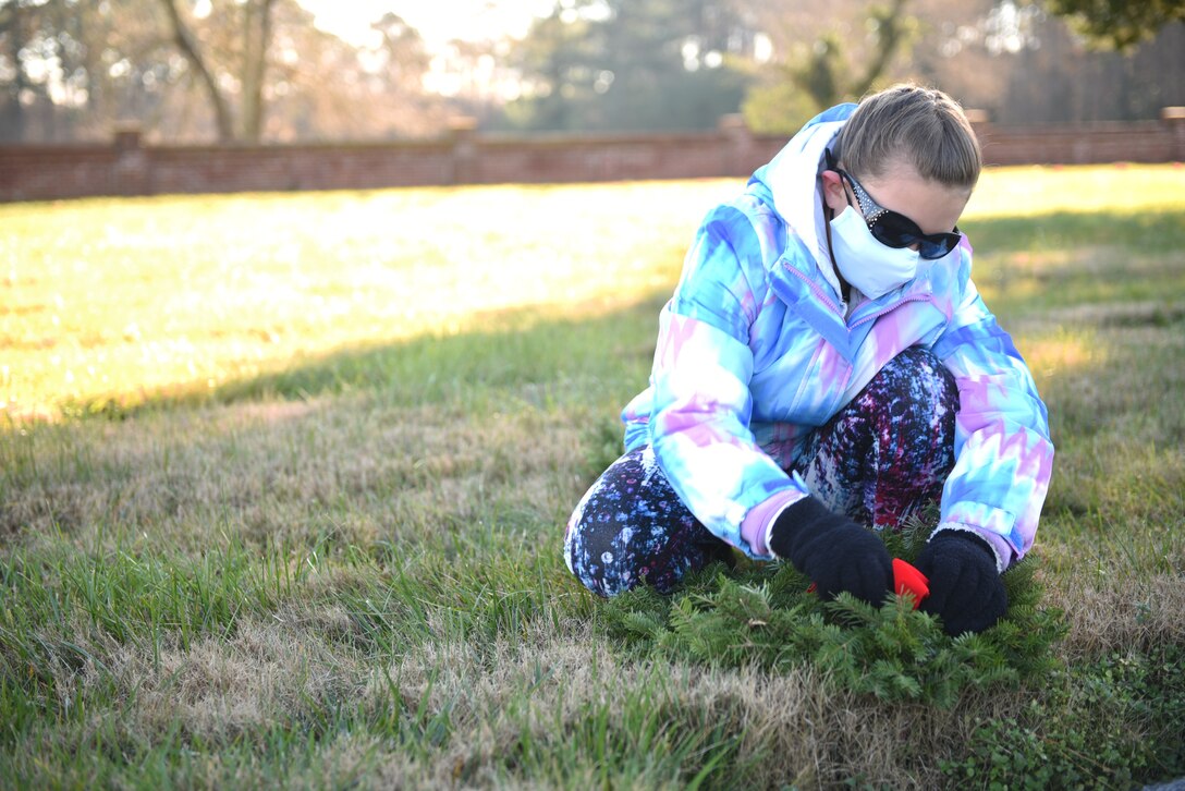 Military child straightens the bow on a wreath covering a headstone.