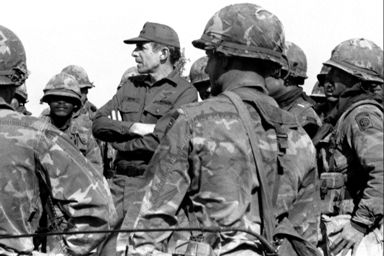 A man wearing a military uniform stands elevated as he talks to soldiers.