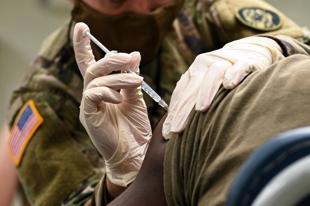 A person wearing a face mask and gloves gives an injection to a soldier.