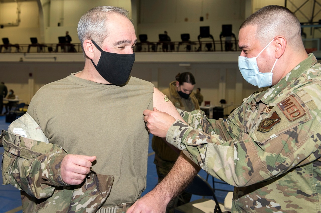 A man in a military uniform applies a small band aid to the arm of another man in military uniform.