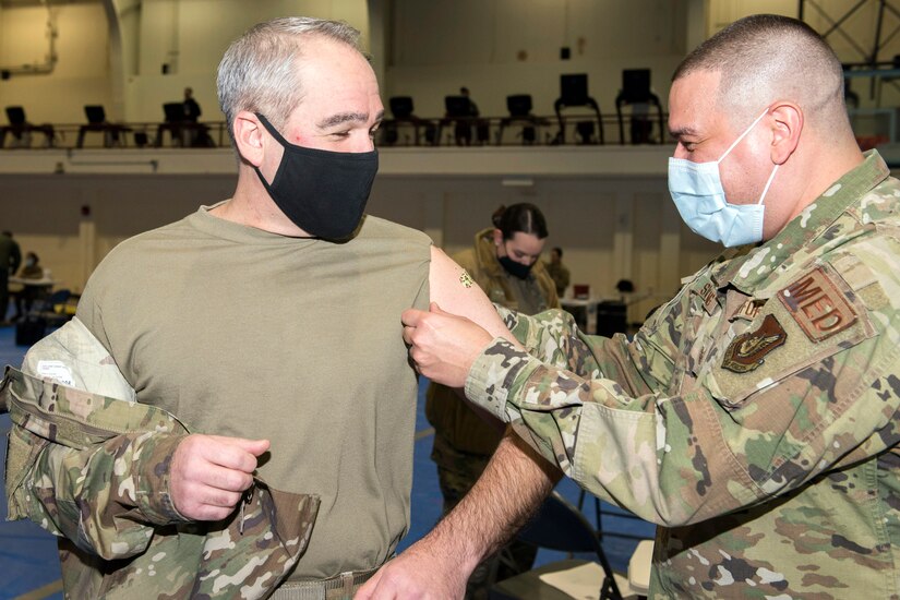 A man in a military uniform applies a small band aid to the arm of another man in military uniform.