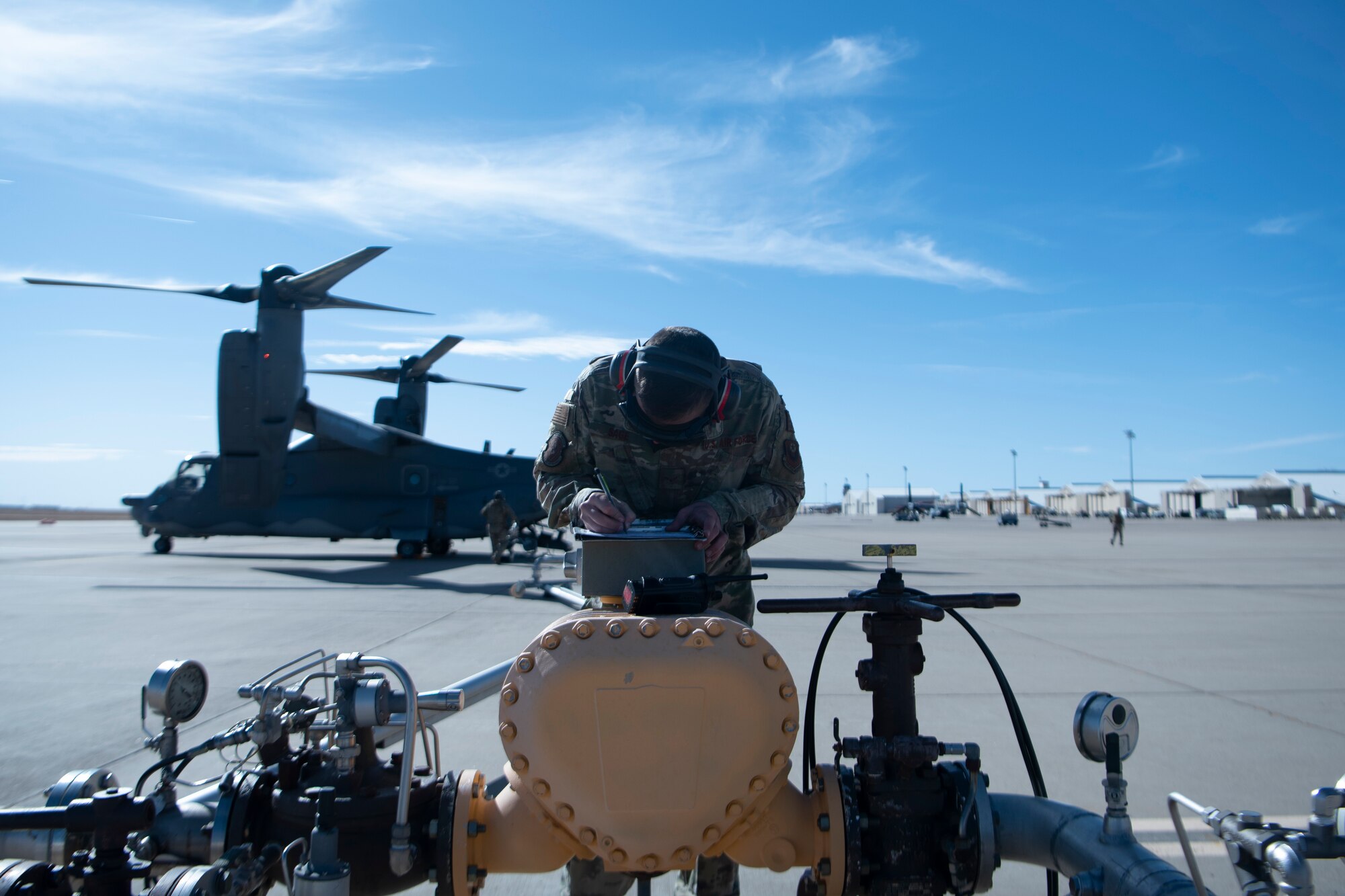 An Airman stands center screen over fuel pipes while taking notes regarding the fuel