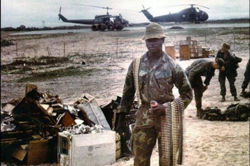 A man in fatigues poses with bands of ammunition as two helicopters and other service members mill around in the background.