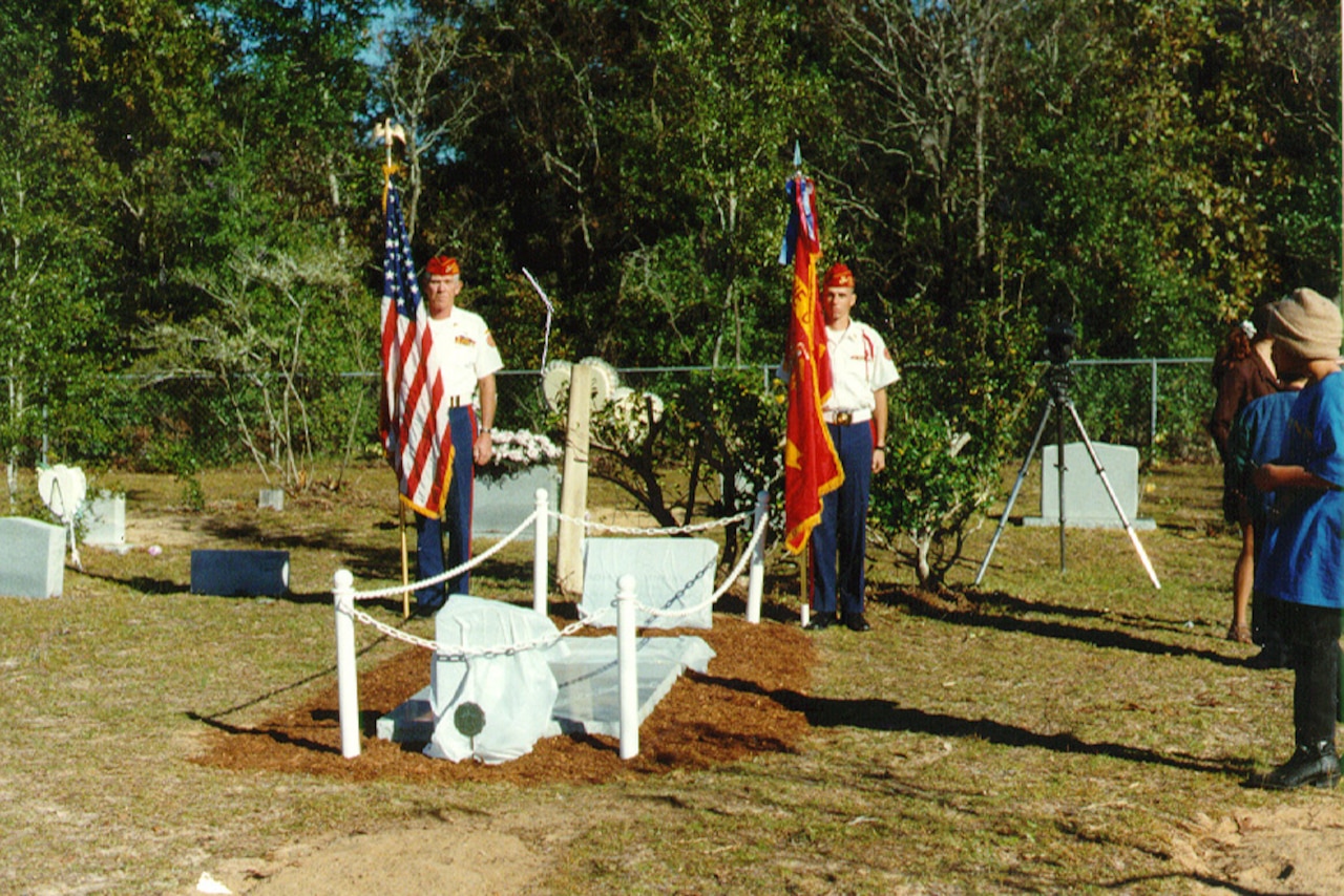 Two Marines hold flags beside a grave cordoned off with a small white chain-link fence.