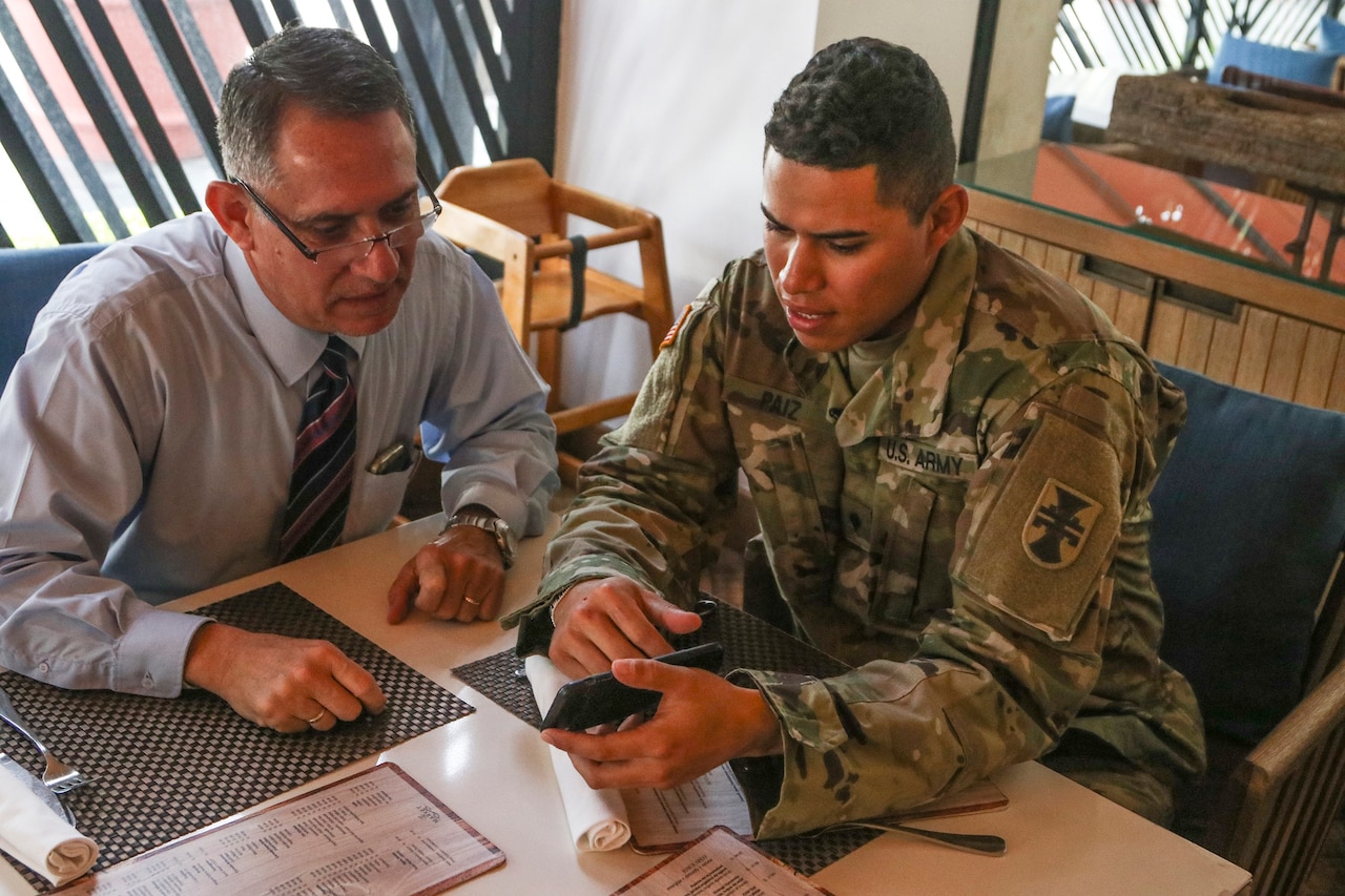A soldier shares a cell phone image with another man.