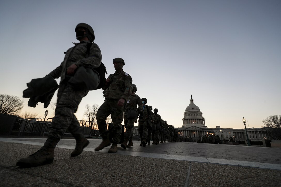 National Guard soldiers and airman walk in a single-file line on pavement, with the Capitol dome in the background.