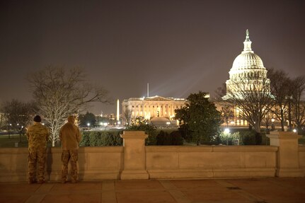Two soldiers stand guard at night with the Capitol building lit up in the background.