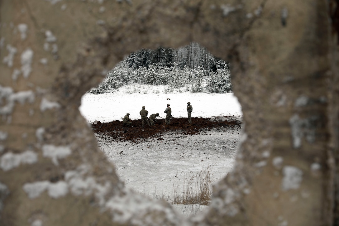 Soldiers seen through a hole in a cement wall work in a field.