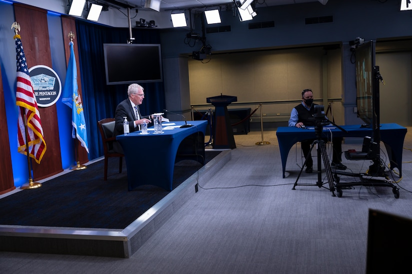 A man seated at a table with  microphone, speaks on a video screen.