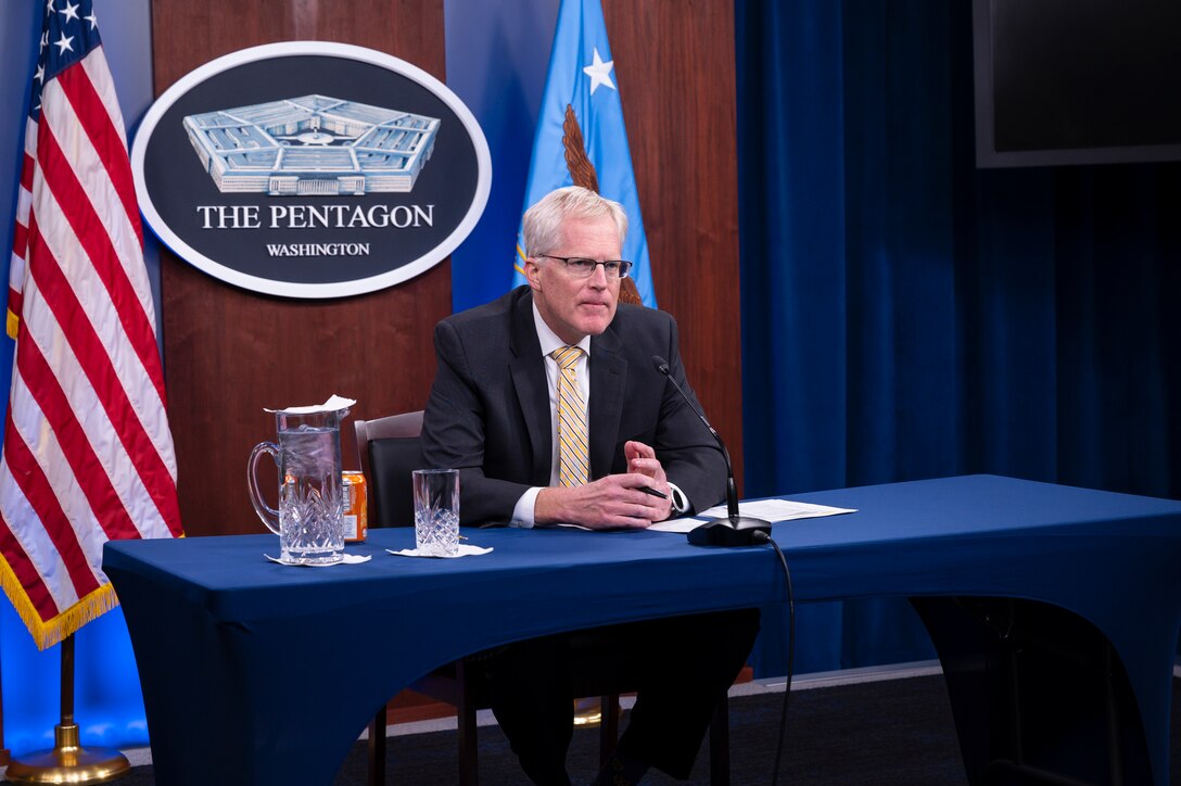 A man sits at desk and speaks into a microphone. A plaque saying “The Pentagon” and two flags are behind him.