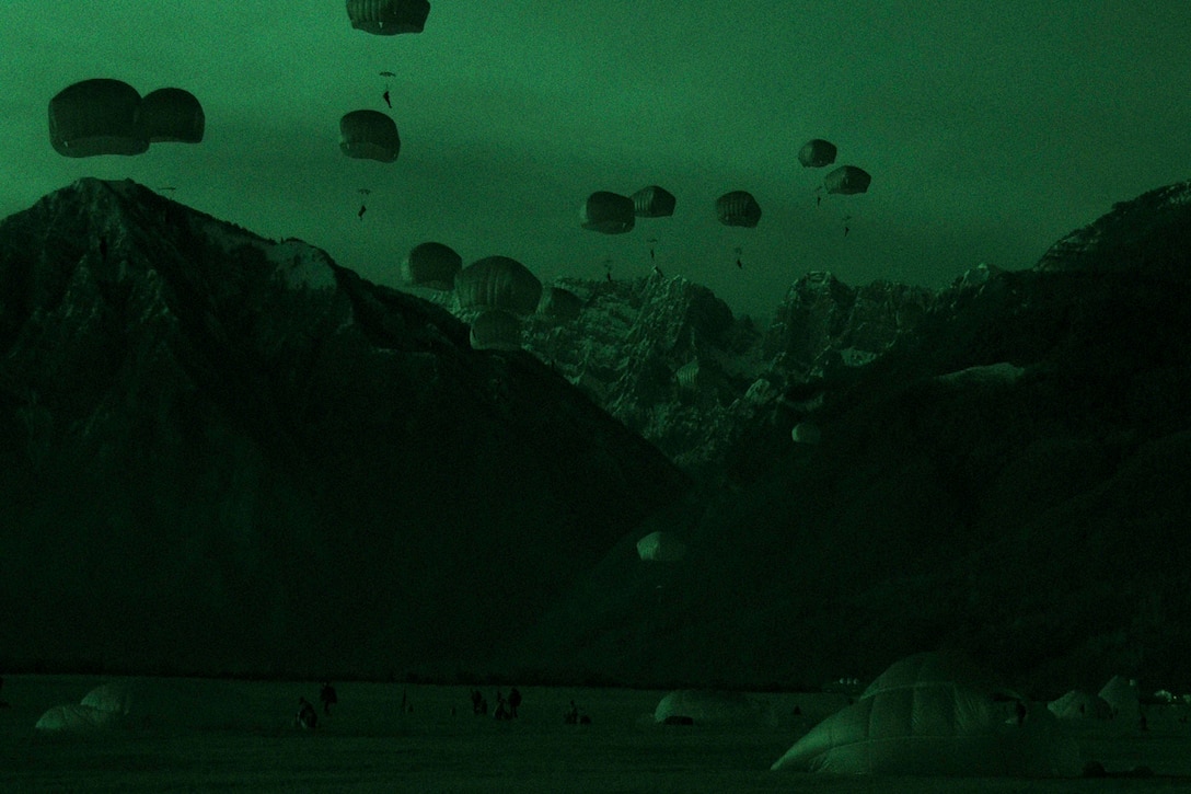 Soldiers freefall wearing parachutes near mountains.