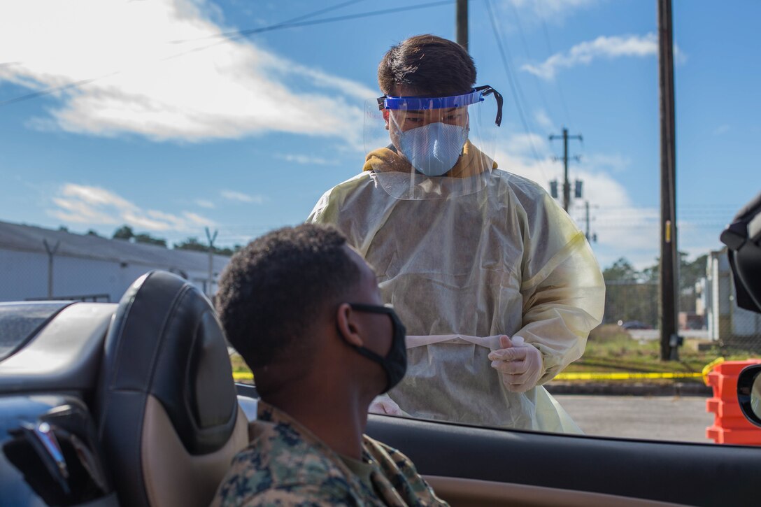 A Navy seaman wearing personal protective equipment stands on the driver's side of a vehicle and talks to a service member who is sitting in his vehicle.