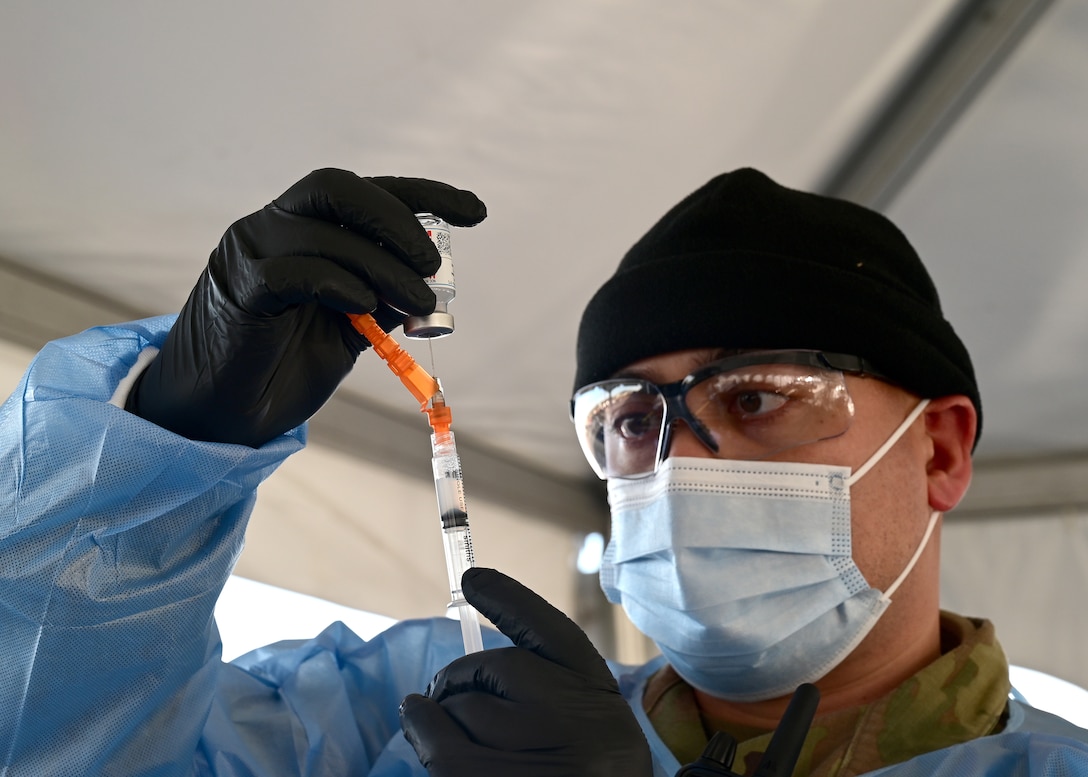 A soldier wearing a face mask and gloves uses a syringe to draw liquid from a small bottle.