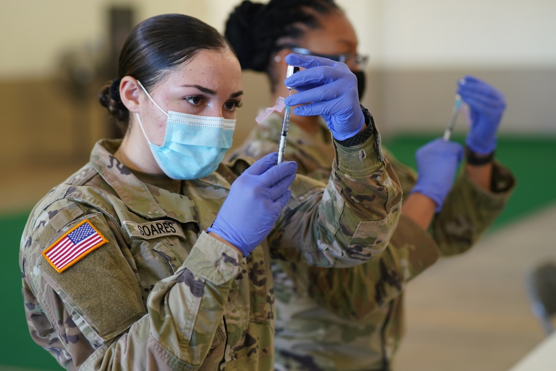 Two soldiers wearing face masks and gloves use syringes to draw medicine from a small bottle.
