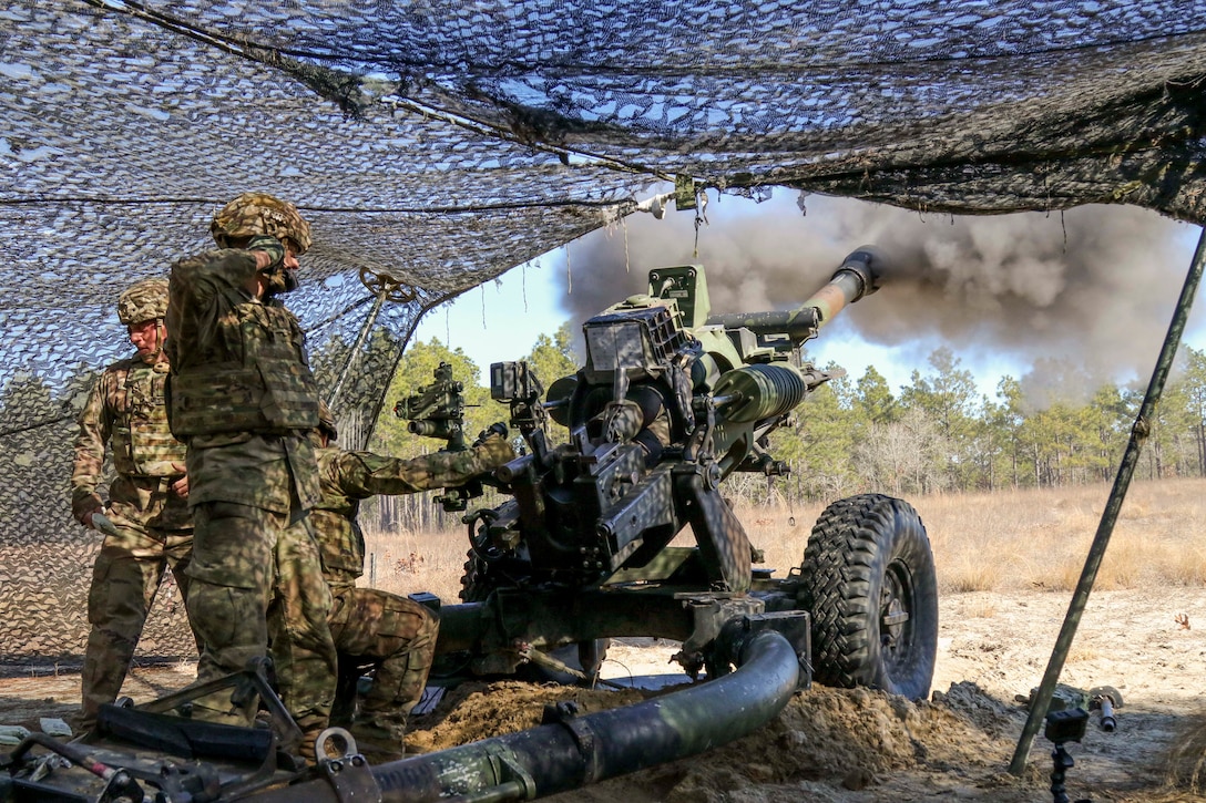 Soldiers fire a weapon under a netted tent.