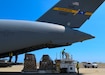 Two people stand on aircraft loading equipment, one above and one behind, ready to load the airplane from the rear. A view of the large rear section of a C-17 is in the shot as well.