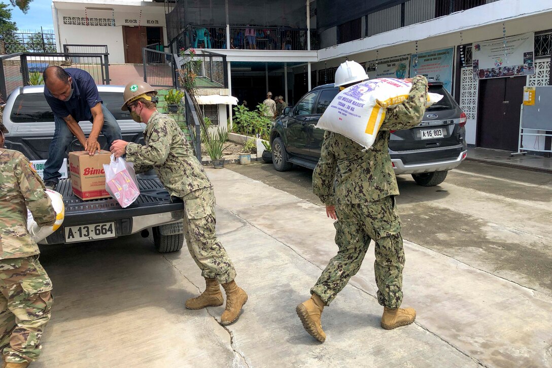Seabees unload and  carry large bags of food across a street.