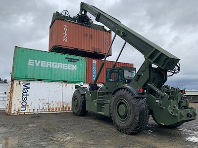 A larger Rough Terrain Container Handler stacks a shipping container on the third level of a stack of other shipping containers in a training exercise.