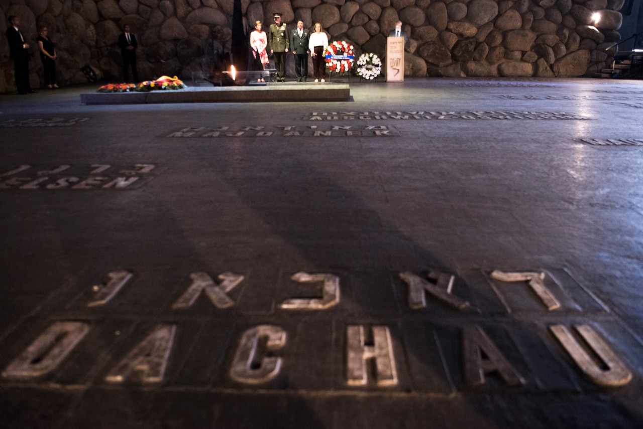 Two men dressed in military uniforms and two women stand beside two wreaths and a flame at a memorial.