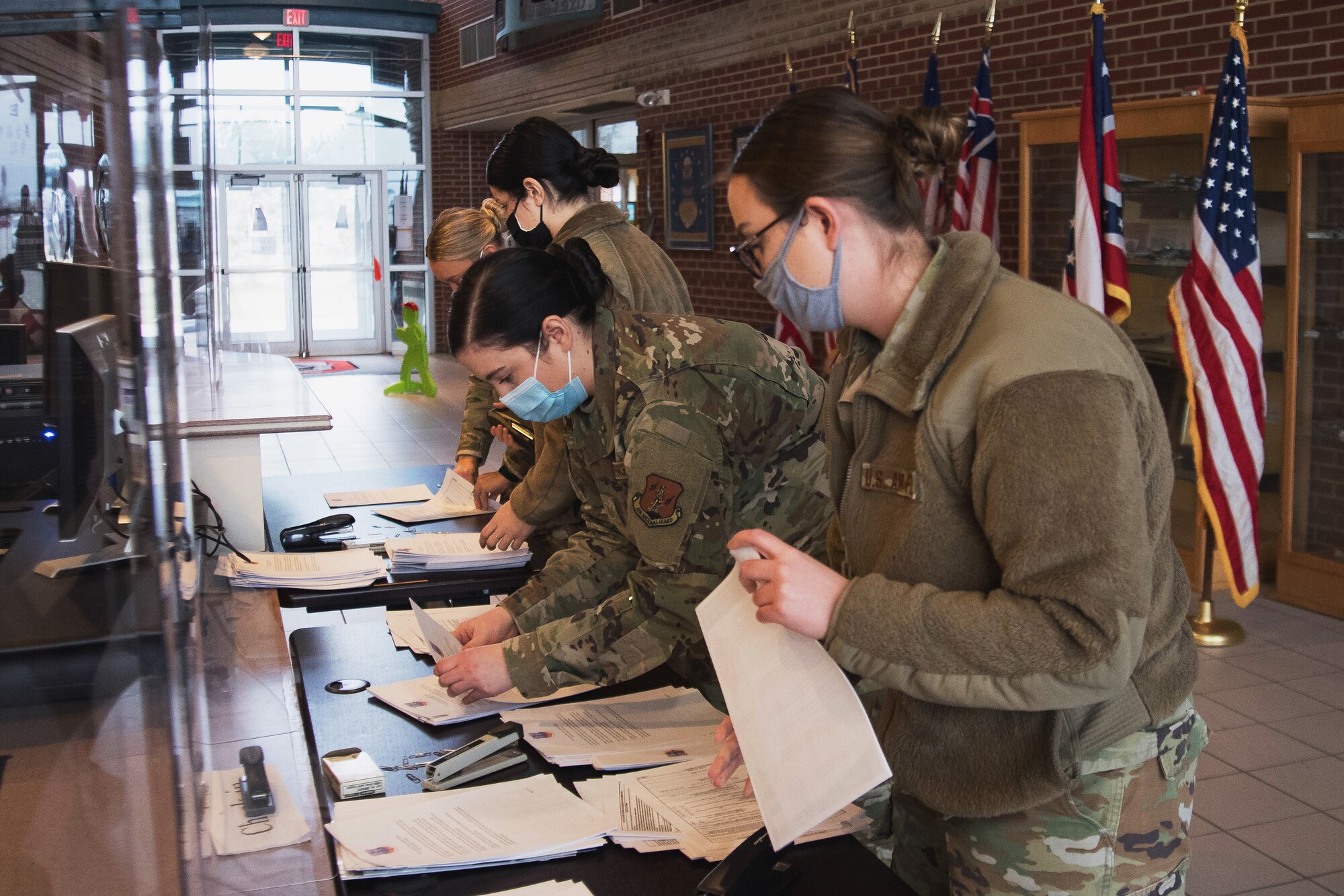 Four Airmen prepare paperwork on a table for a processing line for administration of the COVID-19 vaccination,