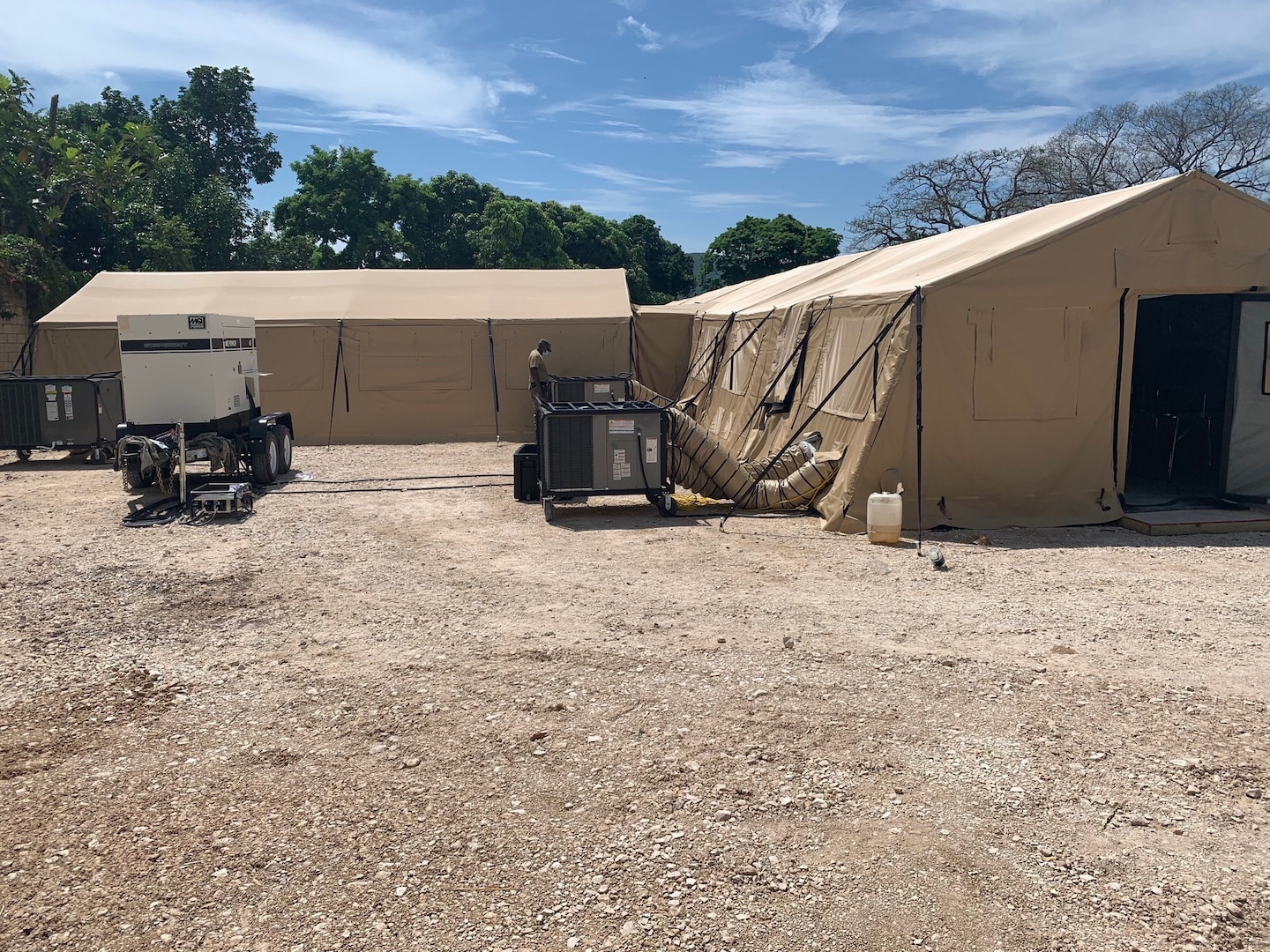 Two tan tent structures with support equipment outside are set up in a dirt field.