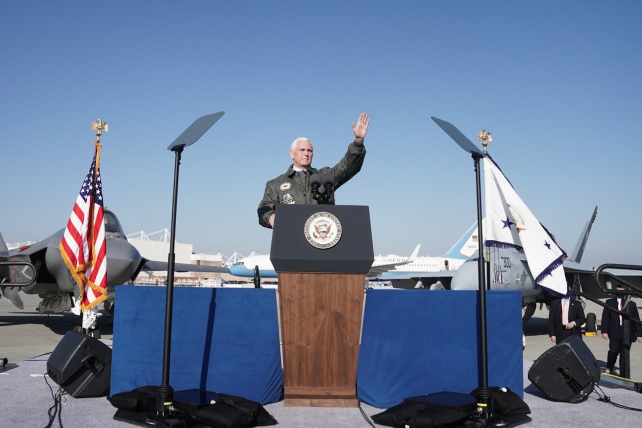 A man wearing a light jacket stands under a sunny sky and waves to an audience assembled on an airfield; a number of planes are in the background.