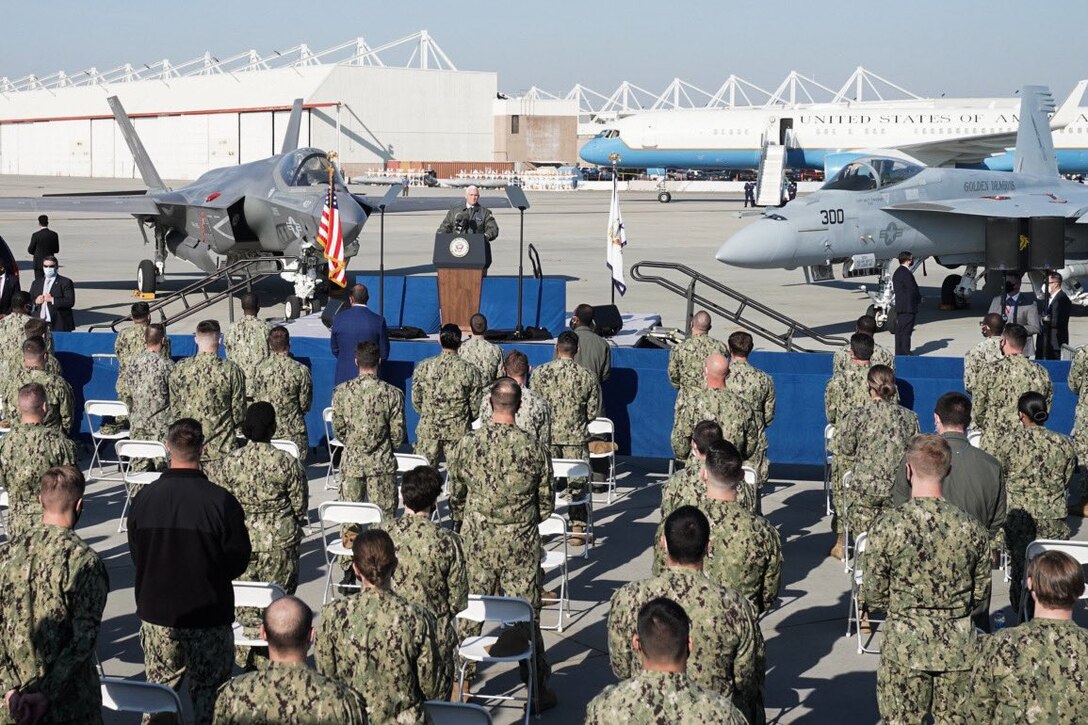 Service members stand beside their chairs on an airfield as a man standing on a stage in front of them speaks into a microphone.