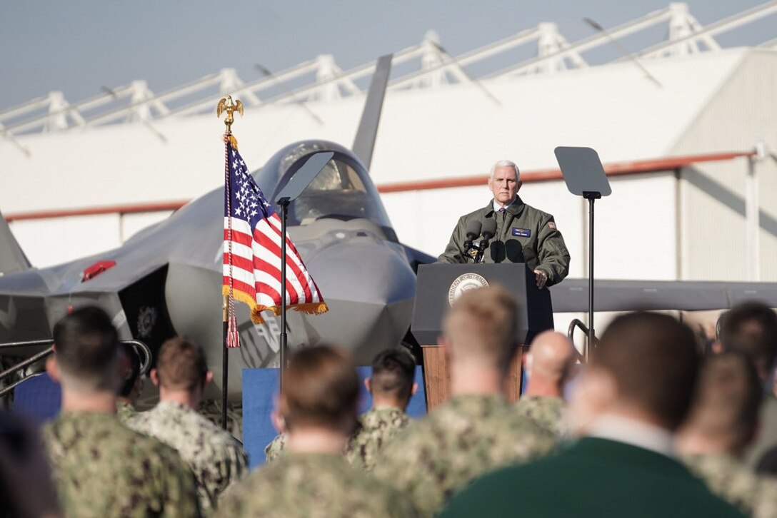 A U.S. flag waves in the breeze as a man stands behind a podium to speak; a jet is parked behind him.